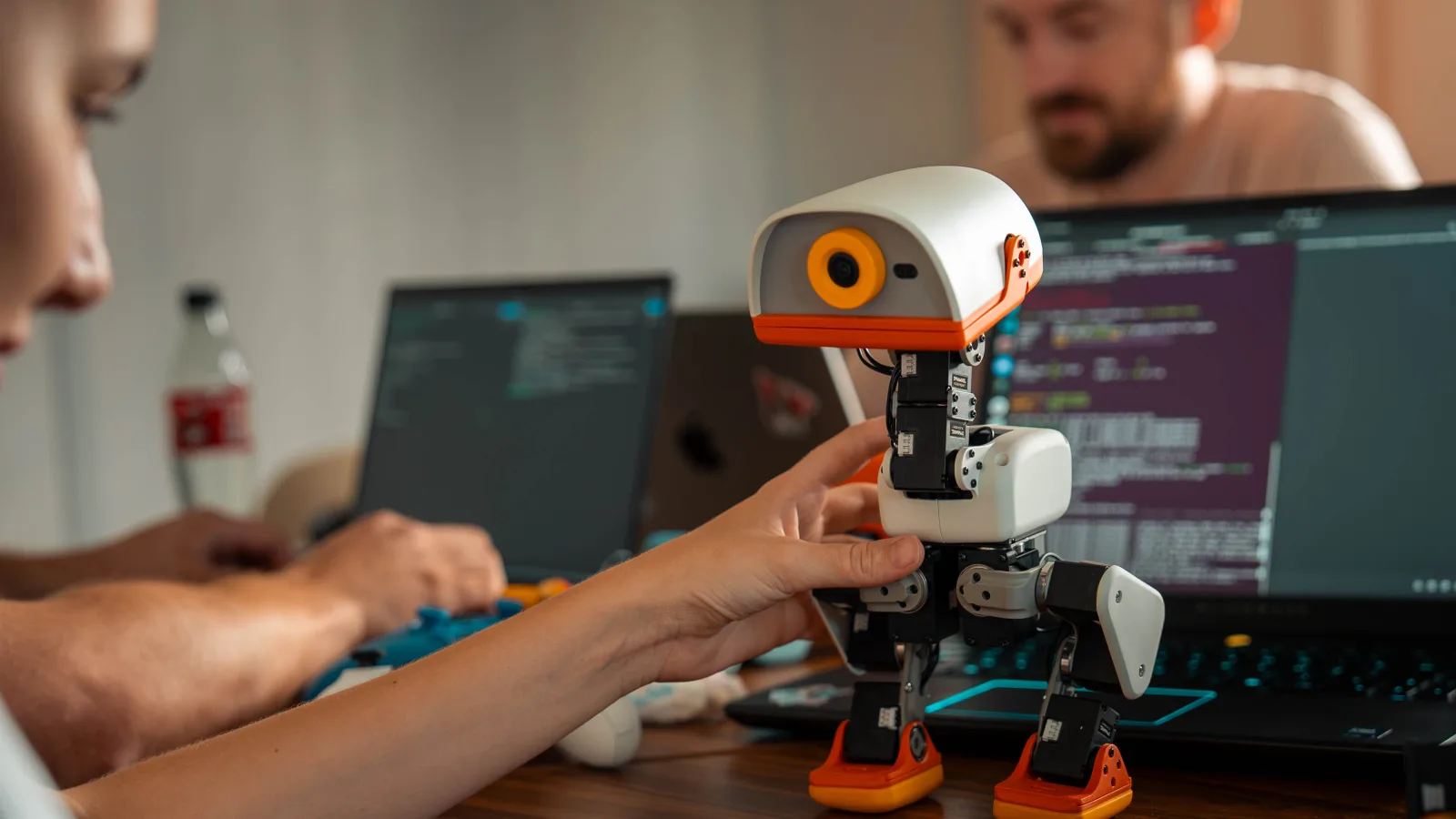 A white-and-orange Microduck robot on a desk between two laptops.